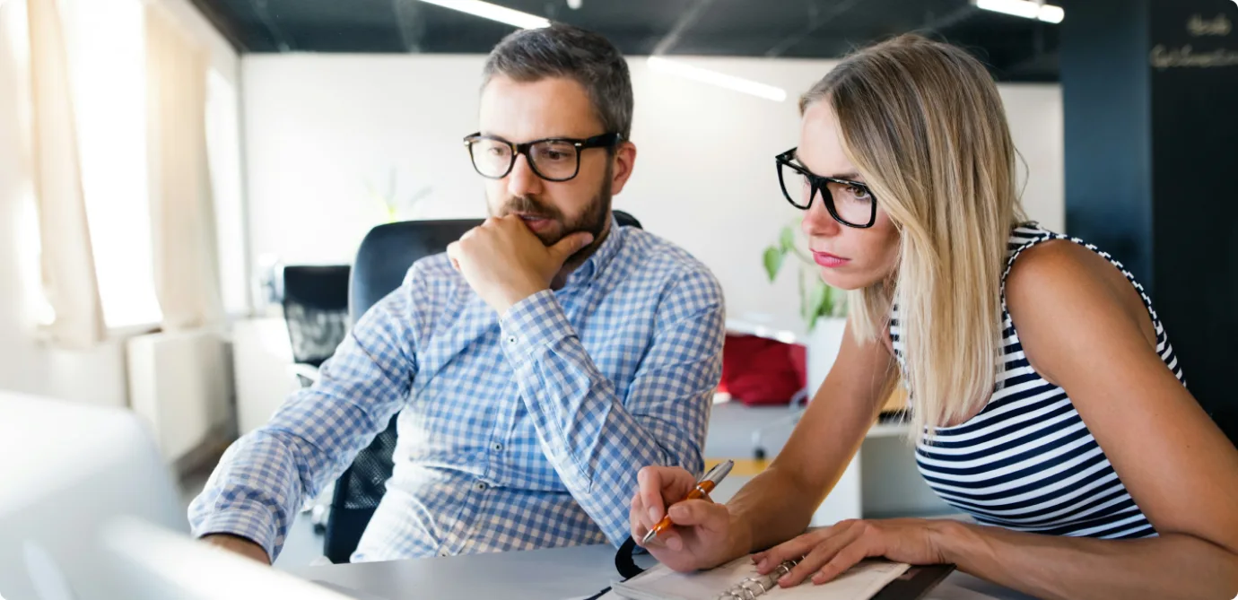 Two colleagues in glasses reviewing notes together at a desk